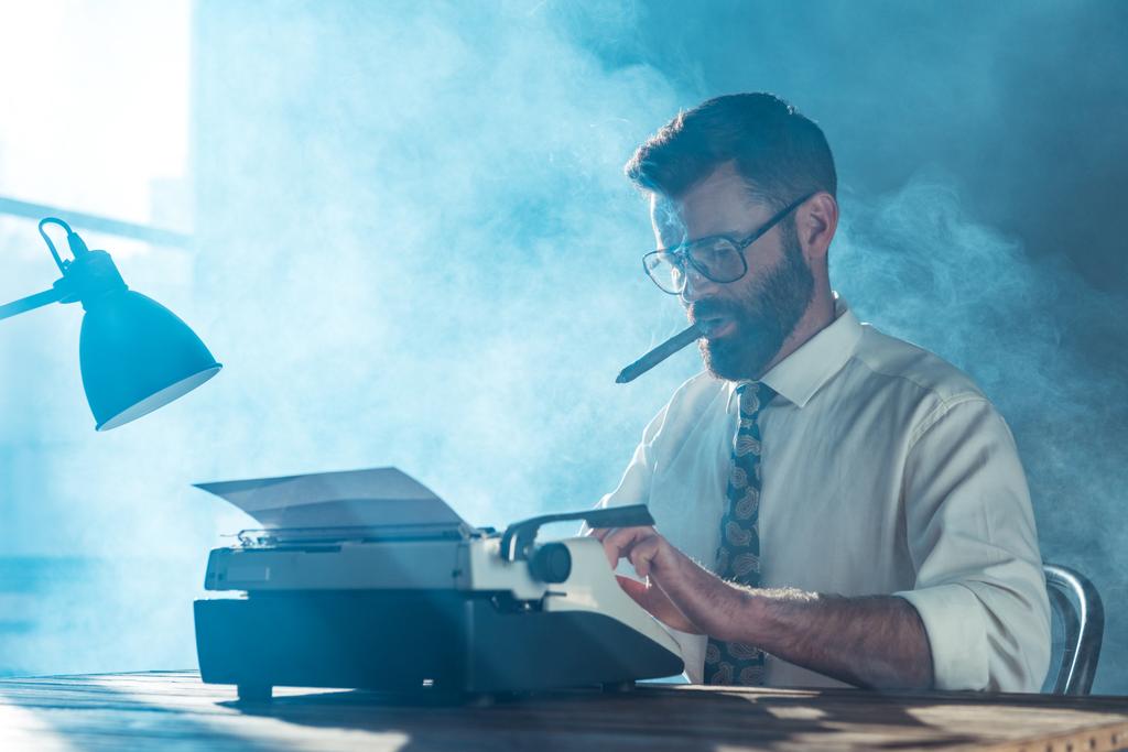 jornalista barbudo em óculos sentado à mesa, digitando em máquina de escrever vintage e fumar perto da janela
 - Foto, Imagem