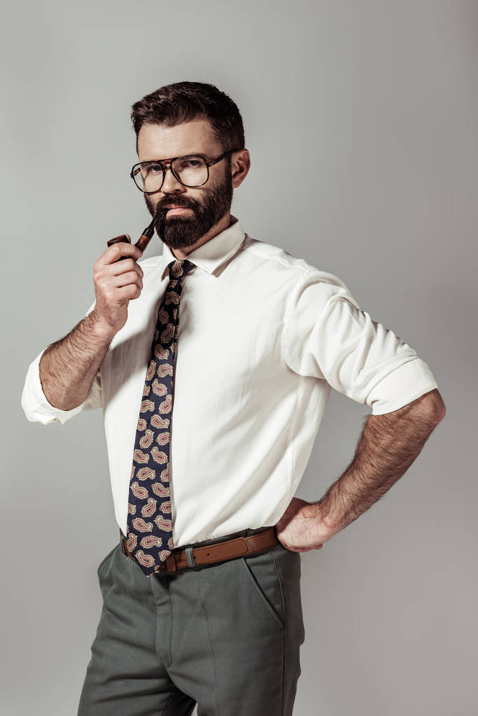 όμορφος γενειοφόρος άνδρας με γυαλιά και πουκάμισο σωλήνα το κάπνισμα και να βλέπουν τα φωτογραφικών μηχανών που απομονώνονται σε γκρι - Φωτογραφία, εικόνα