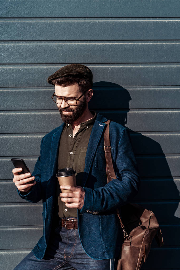 красивий бородатий чоловік в окулярах і кепці тримає паперову чашку і використовує смартфон
 - Фото, зображення