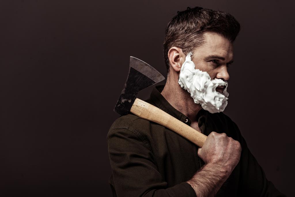 handsome bearded man with shaving foam on face holding ax isolated on brown - Photo, Image