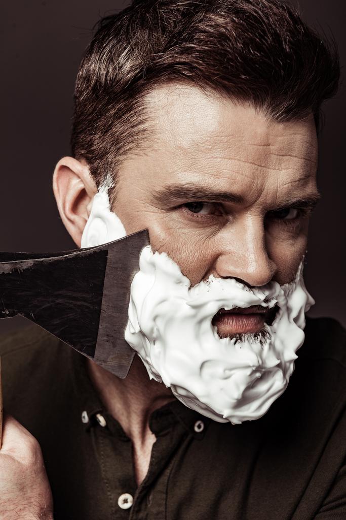 close up of serious handsome bearded man shaving with ax while looking at camera isolated on brown - Photo, Image