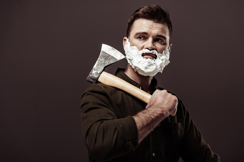 красивый бородатый мужчина с пеной для бритья на лице держа топор изолирован на коричневый
 - Фото, изображение