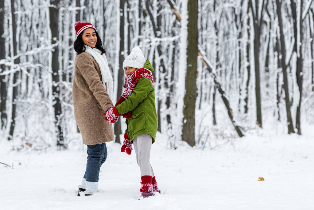 african american matka i Córka, uśmiechając się, trzymając się za ręce i patrząc na kamery w winter park - Zdjęcie, obraz