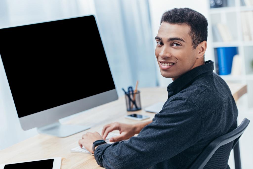 biznesmen młodych mieszanej rasy przy użyciu komputera stacjonarnego z pustego ekranu i uśmiecha się do kamery  - Zdjęcie, obraz