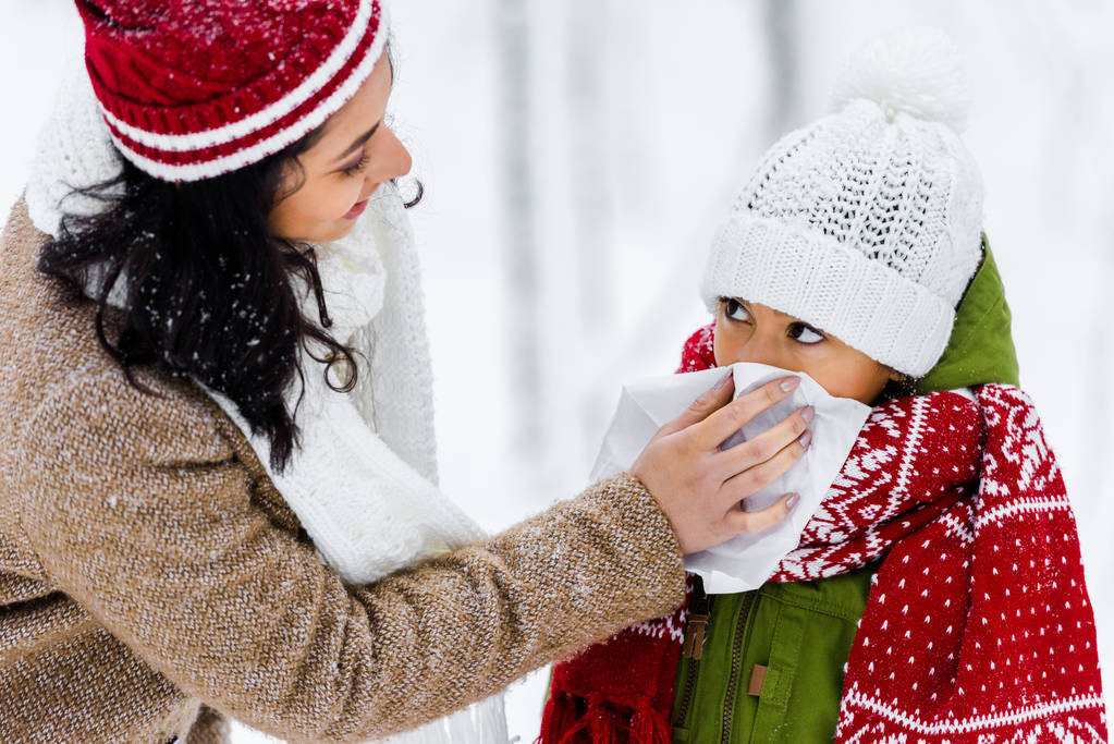 african american woman wiping with napkin nose of cute daughter in winter park  - Photo, Image