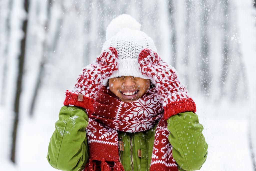 sorrindo criança afro-americana com chapéu de malha puxado sobre os olhos durante a queda de neve no parque de inverno
 - Foto, Imagem