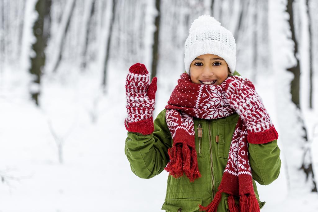 Afrikanisch-amerikanisches Kind in warmer Kleidung lächelt, winkt mit der Hand und blickt in die Kamera im Winterpark - Foto, Bild