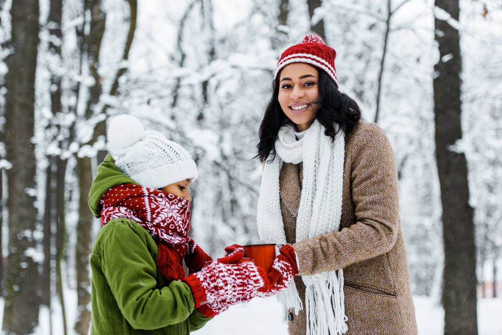 魅力的なアフリカ系アメリカ人の母とお茶のカップを持って、雪に覆われた森でカメラで笑顔のかわいい娘 - 写真・画像