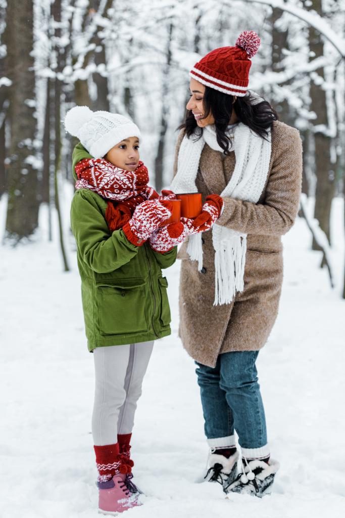 attractive african american mother and cute daughter talking to each other and holding cups of tea in snowy forest - Photo, Image