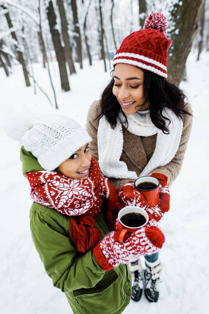 attractive african american mother and cute daughter holding red cups of tea while staying near trees in snowy forest - Photo, Image