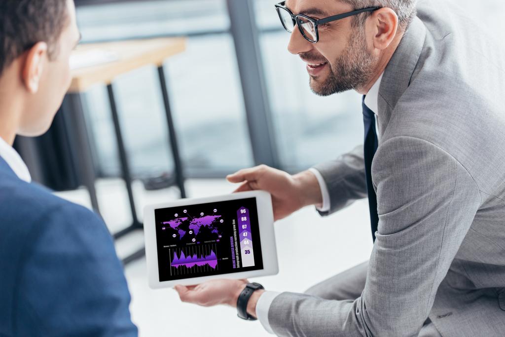 Oříznout záběr podnikatel v brýlích ukazuje digitální tablet s grafy a grafy na obrazovce na mužské kolegy v kanceláři - Fotografie, Obrázek