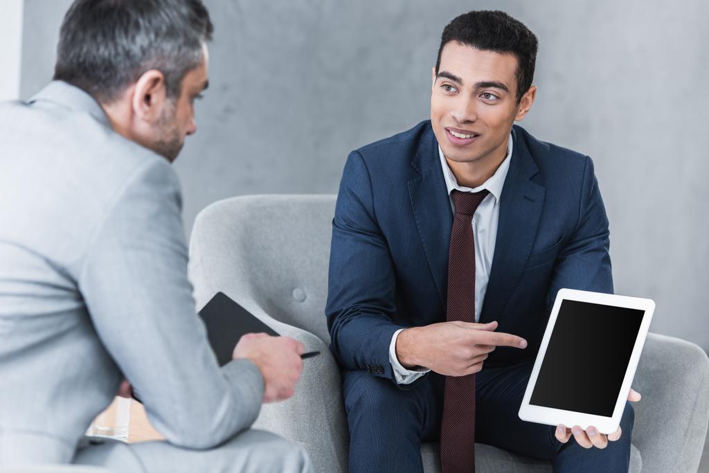 sorridente jovem empresário apontando para tablet digital com tela em branco e olhando para colega masculino durante a conversa
 - Foto, Imagem