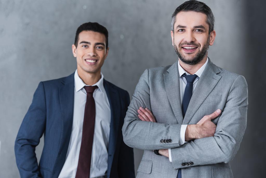 профессиональные уверенные бизнесмены, стоящие вместе и улыбающиеся в камеру на сером
 - Фото, изображение