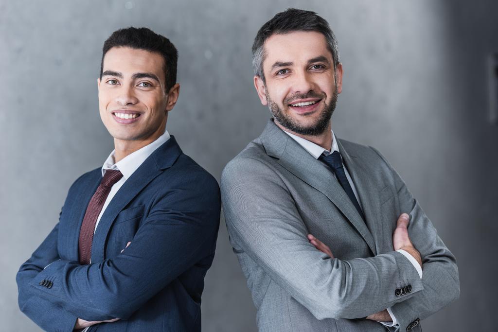 профессиональные уверенные бизнесмены, стоящие со скрещенными руками и улыбающиеся в камеру на сером
 - Фото, изображение