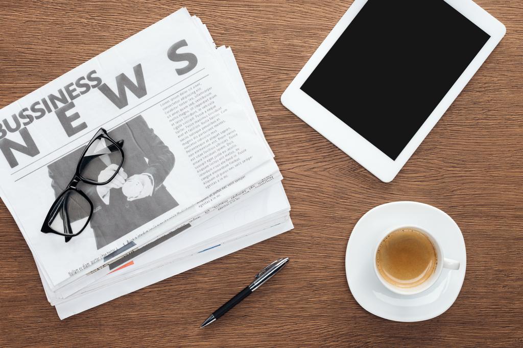 elevated view of cup of coffee, pen, tablet with blank screen and business newspapers on wooden tabletop - Photo, Image