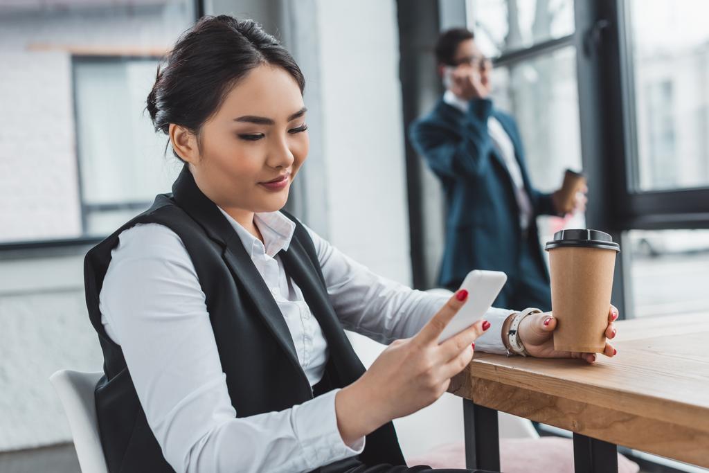 bella donna d'affari kazakh sorridente in possesso di caffè per andare e utilizzando smartphone sul posto di lavoro
 - Foto, immagini
