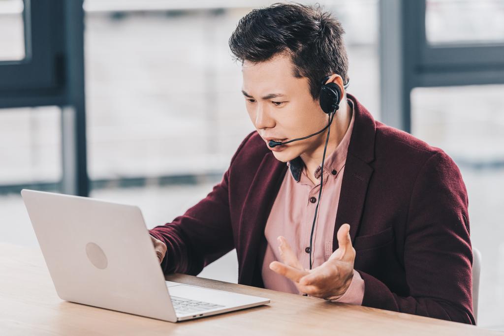 emotionaalinen nuori aasialainen liikemies kuulokkeissa kannettavan tietokoneen avulla työpaikalla
 - Valokuva, kuva
