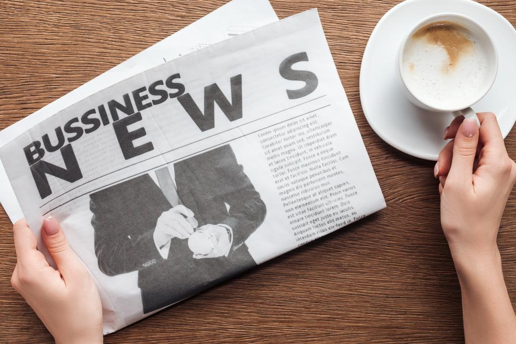 imagen recortada de periodista sosteniendo periódico de negocios y taza de café en la mesa de madera
 - Foto, Imagen