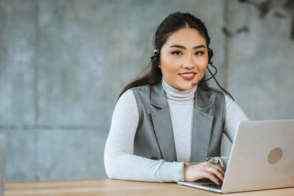 aantrekkelijke jonge zakenvrouw in hoofdtelefoon werken met de laptop en lachend op camera in kantoor - Foto, afbeelding