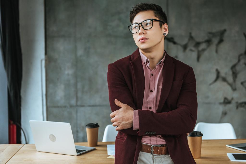 pensivo jovem kazakh empresário em óculos de pé com braços cruzados e olhando para longe no escritório
 - Foto, Imagem