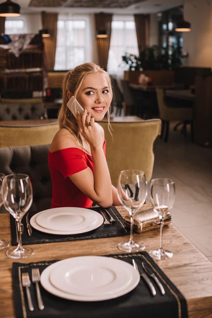 mujer atractiva en vestido rojo sentado en el restaurante con teléfono inteligente y mirando a la cámara
 - Foto, Imagen