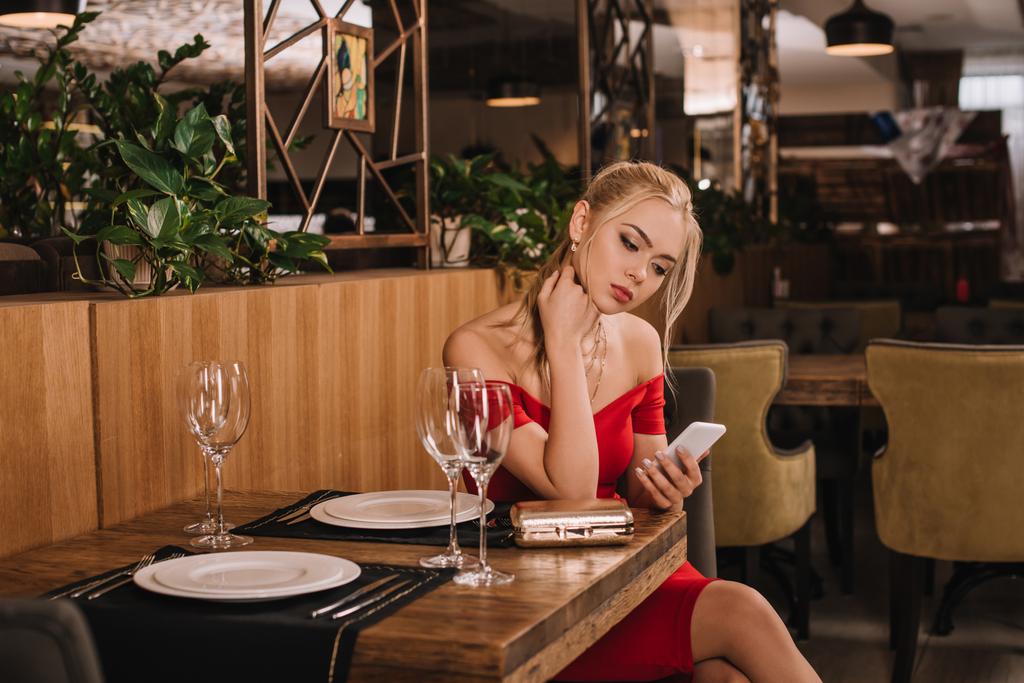 attraktive Frau in rotem Kleid sitzt im Restaurant und schaut aufs Smartphone - Foto, Bild
