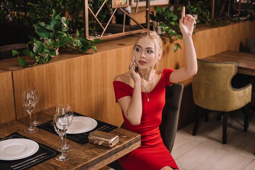 attractive woman in red dress sitting in restaurant and raising hand - Photo, Image