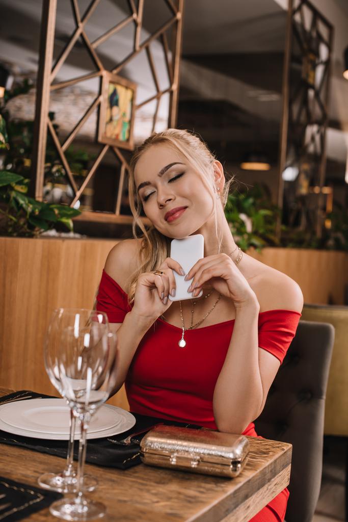 novia de ensueño en vestido rojo sentado en el restaurante y esperando novio con los ojos cerrados
 - Foto, Imagen