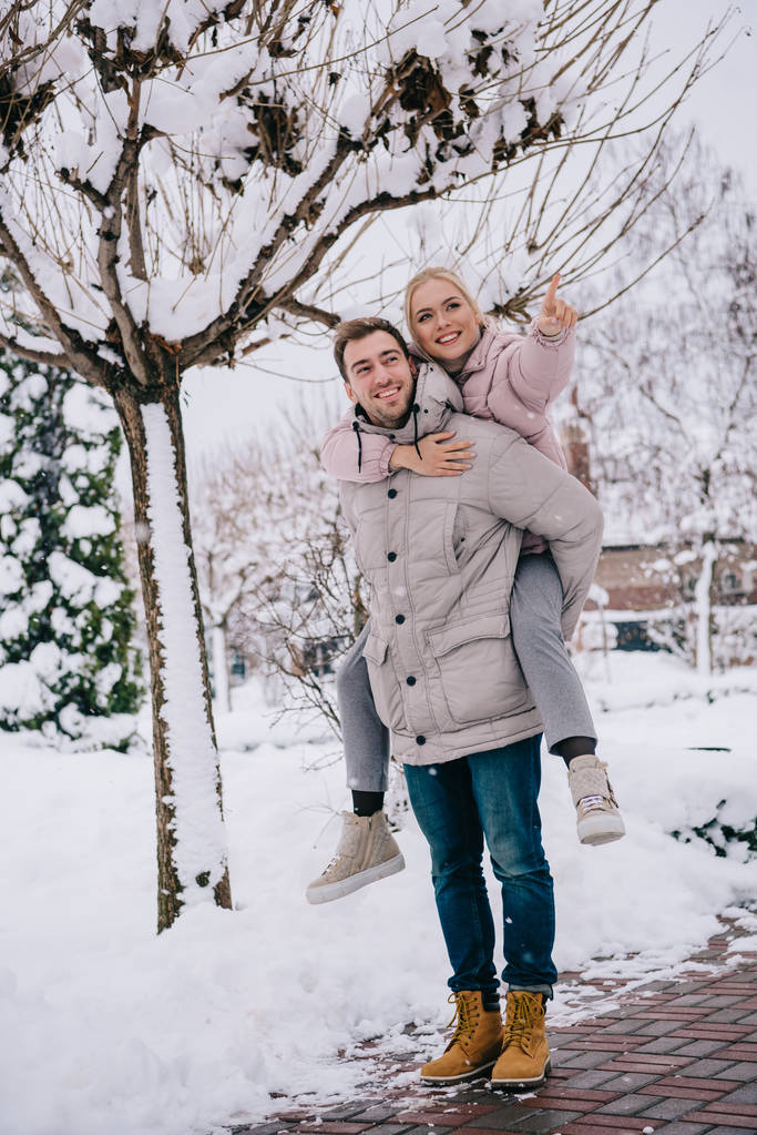 冬に指で指している遊び心のある女性を運ぶハンサムな男 - 写真・画像