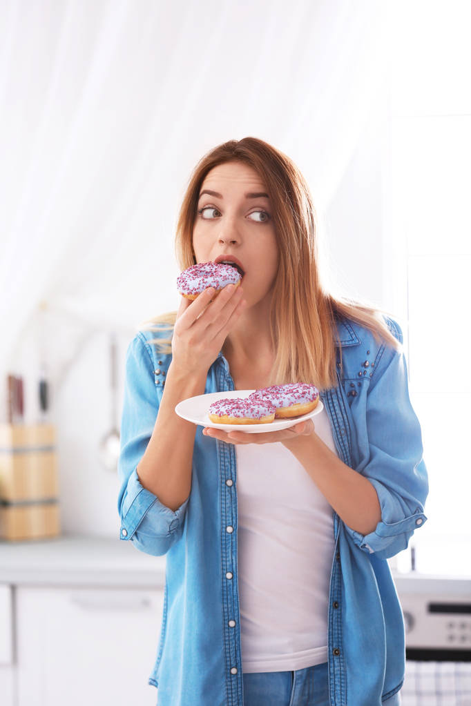 Emocionální mladá žena jíst koblihy v kuchyni. Neúspěšné dieta - Fotografie, Obrázek