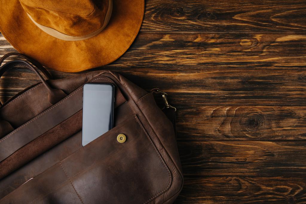 top view of brown leather bag with smartphone and hat on wooden table, travel concept - Photo, Image