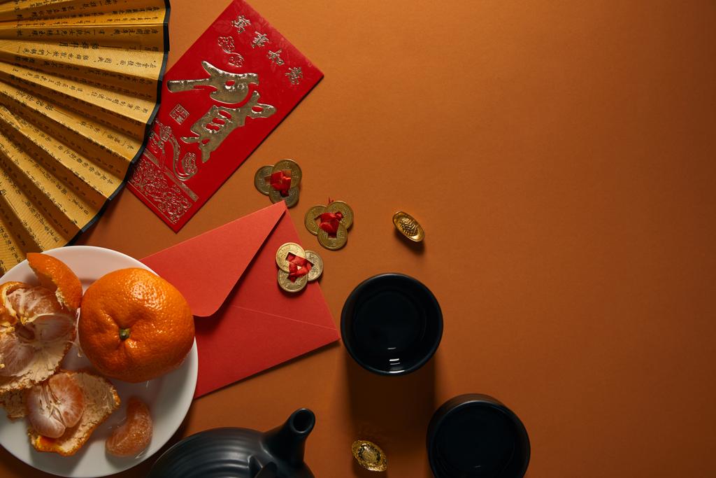 vista dall'alto di mandarini su piatto, set da tè nero, ventaglio con geroglifici, decorazioni dorate e busta rossa su sfondo marrone
 - Foto, immagini