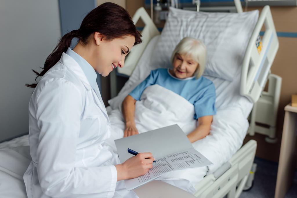 χαμογελαστή γυναίκα γιατρό κάθεται στο κρεβάτι και το γράψιμο διάγνωση ενώ ανώτερος γυναίκα ξαπλωμένη στο κρεβάτι στο νοσοκομείο - Φωτογραφία, εικόνα