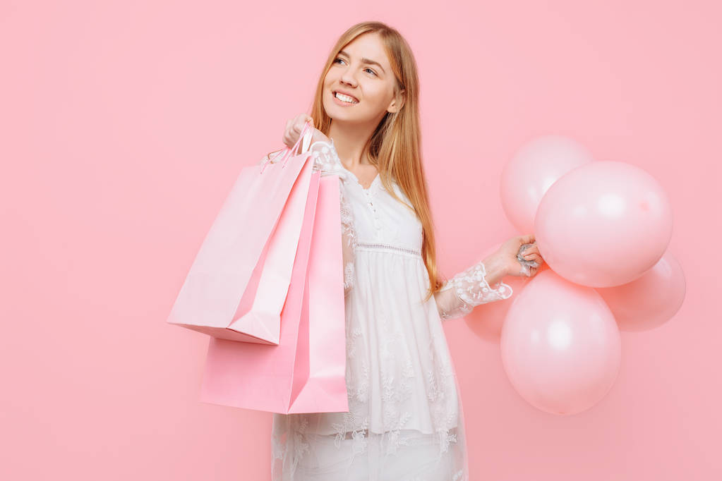 Ευτυχισμένη γυναίκα, σε ένα λευκό φόρεμα, με σακούλες στα χέρια και τα μπαλόνια, σε ροζ φόντο - Φωτογραφία, εικόνα