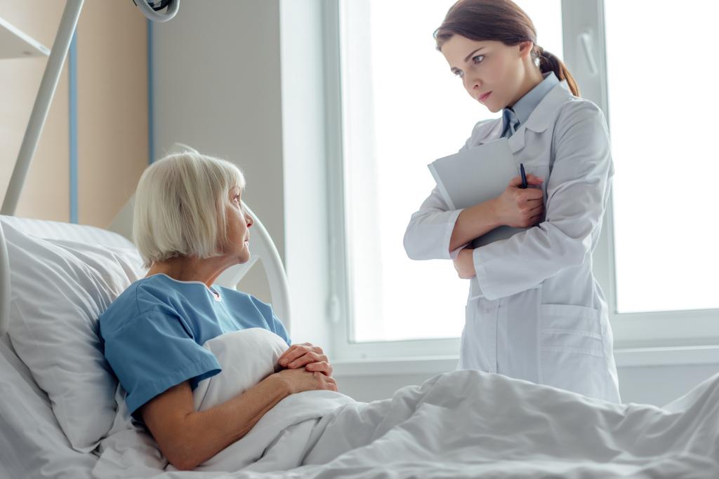 női orvos gazdaság diagnózis, és meglátogatta a kórházi ágyon fekve idősebb nő - Fotó, kép