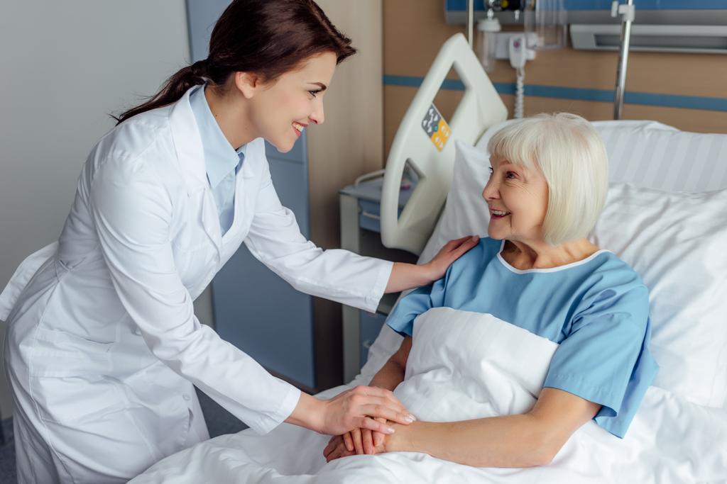 Szczęśliwa kobieta lekarz trzymając się za ręce i konsultacji starszy kobietę leżącą w łóżku szpitalnym - Zdjęcie, obraz