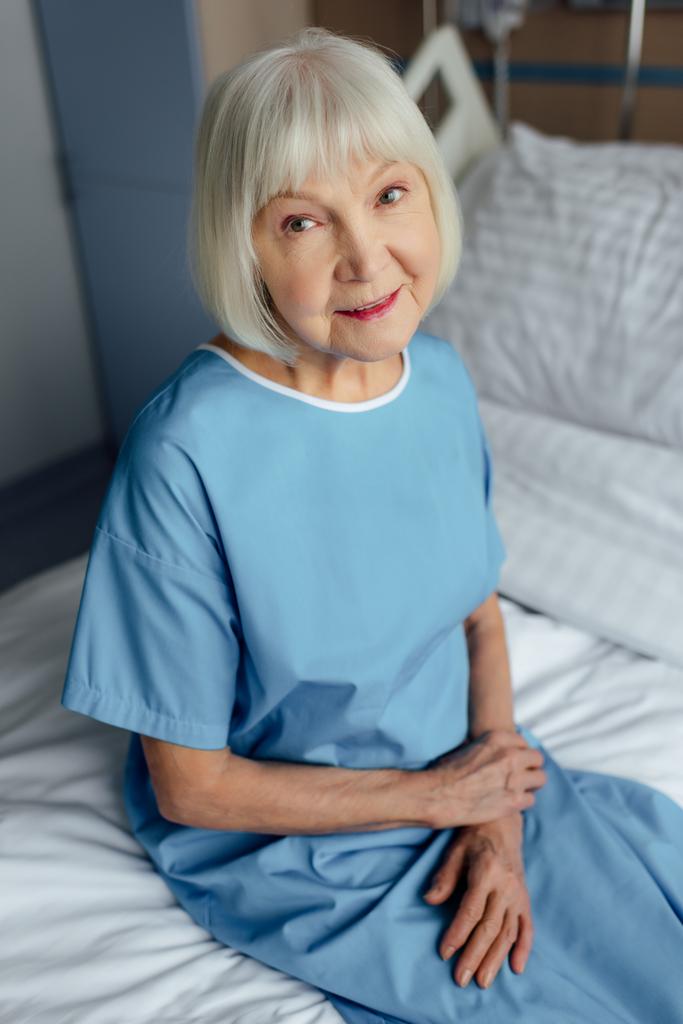 usměvavá starší žena se sepjatýma rukama, sedí na posteli a při pohledu na fotoaparát v nemocnici - Fotografie, Obrázek