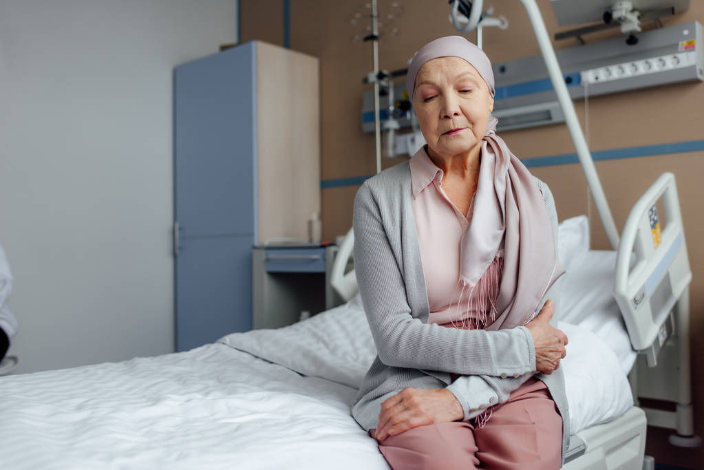 Krebskranke Seniorin sitzt mit gekreuzten Händen auf Bett im Krankenhaus - Foto, Bild