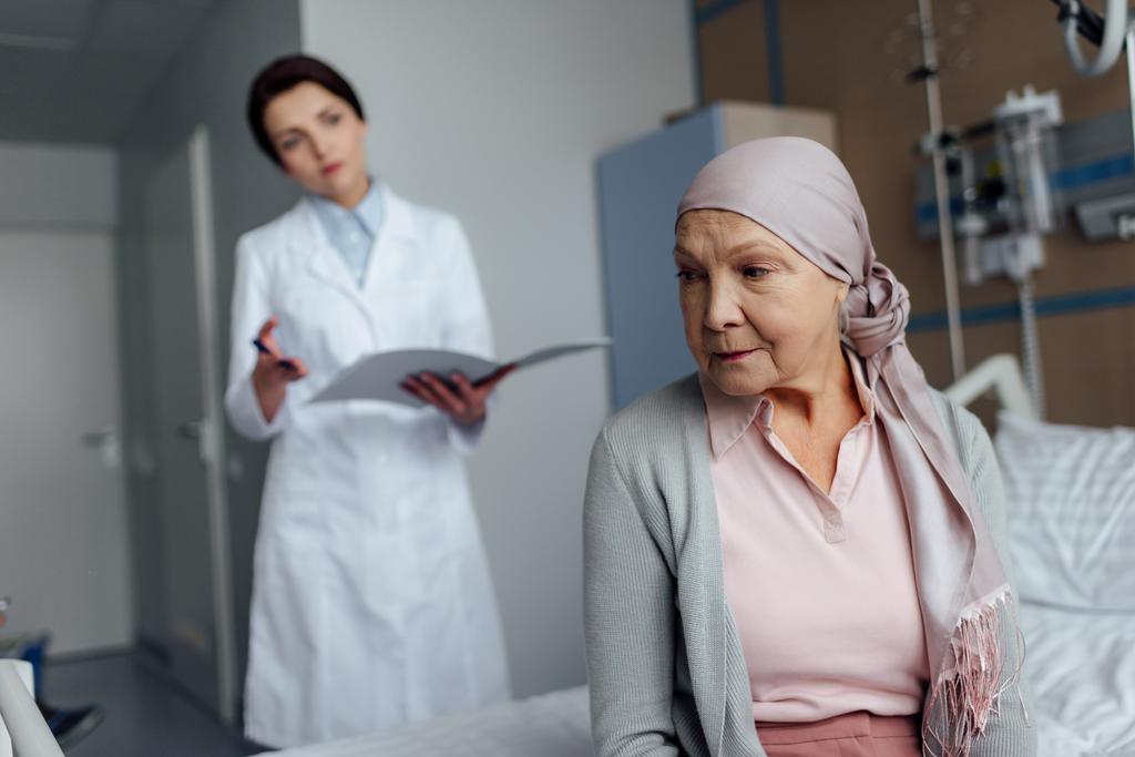 disgustada mujer mayor en pañuelo con cáncer sentada en la cama del hospital con el diagnóstico de la mujer en segundo plano
 - Foto, Imagen