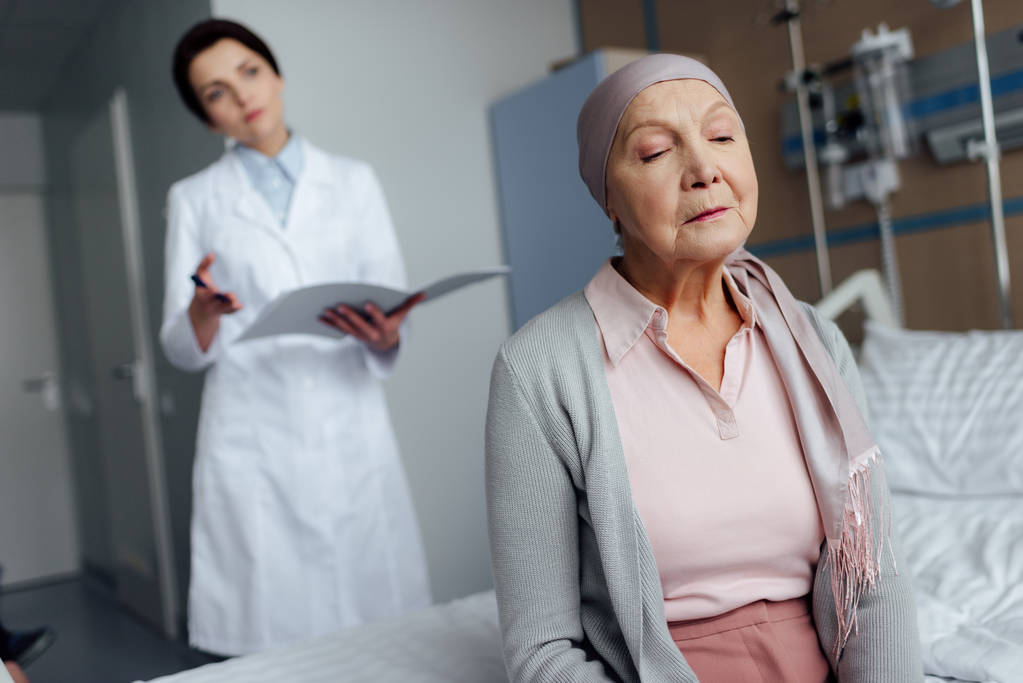 mujer mayor triste en el pañuelo con cáncer sentado en la cama del hospital con médico femenino que sostiene el diagnóstico en el fondo
 - Foto, Imagen