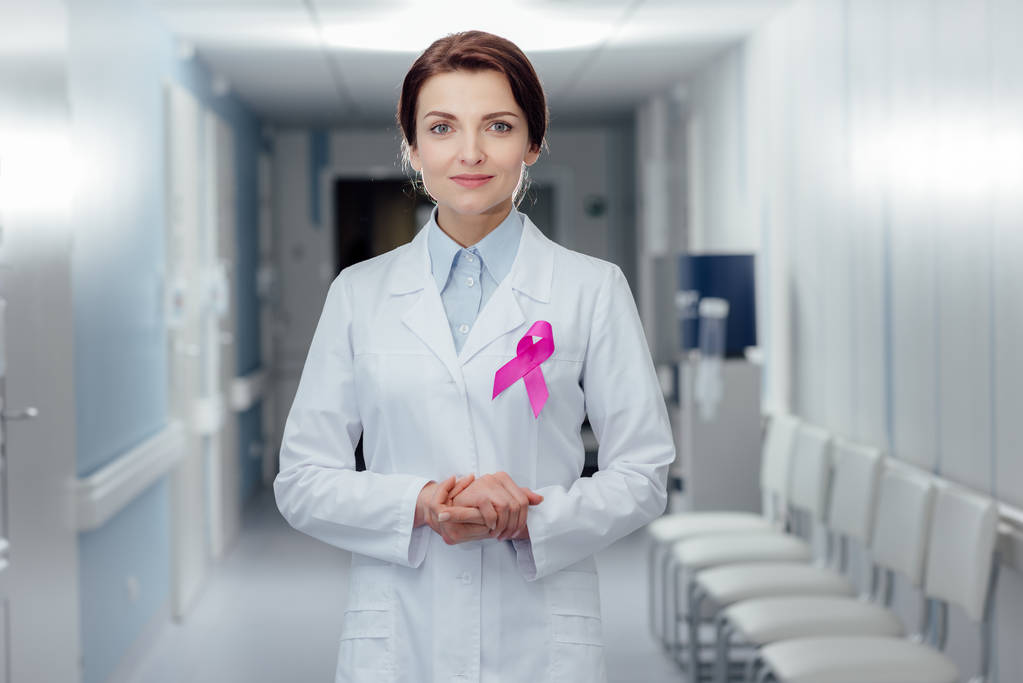 femme médecin avec ruban rose et les mains pliées regardant la caméra à l'hôpital, concept de sensibilisation au cancer du sein
 - Photo, image