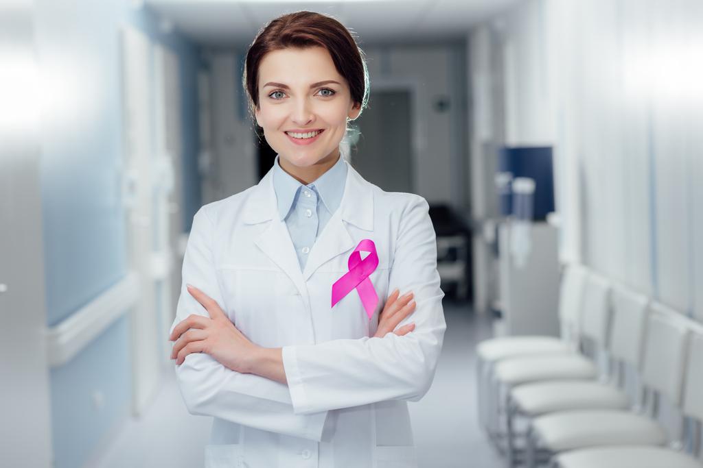 smiling female doctor with pink ribbon and arms crossed in hospital, breast cancer awareness concept - Photo, Image