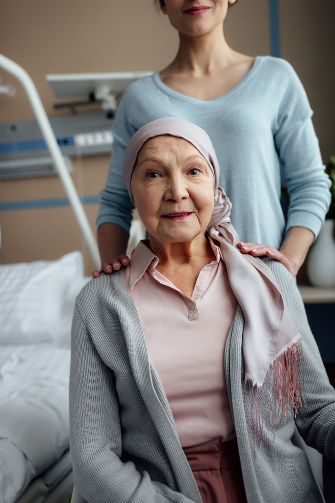 smiling senior woman in kerchief looking at camera with daughter on background in hospital - Photo, Image