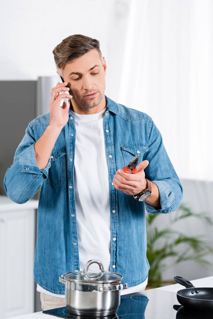 красивый мужчина разговаривает на смартфоне и держит плоскогубцы в руке, стоя на кухне
  - Фото, изображение