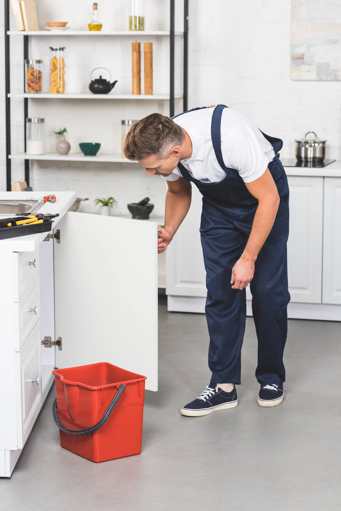 Aikuinen mies työvaatteissa korjaamassa keittiön pesuallasta
  - Valokuva, kuva