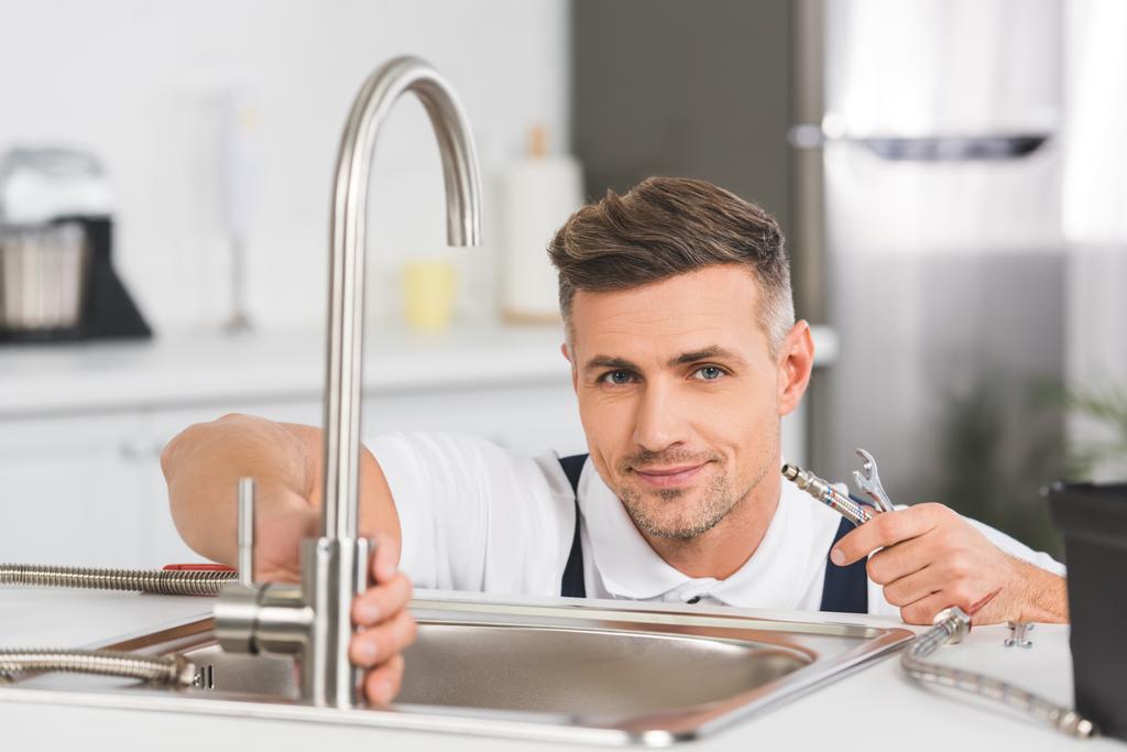 smiling adult repairman holding pipe and spanner while repairing faucet at kitchen and looking at camera - Photo, Image