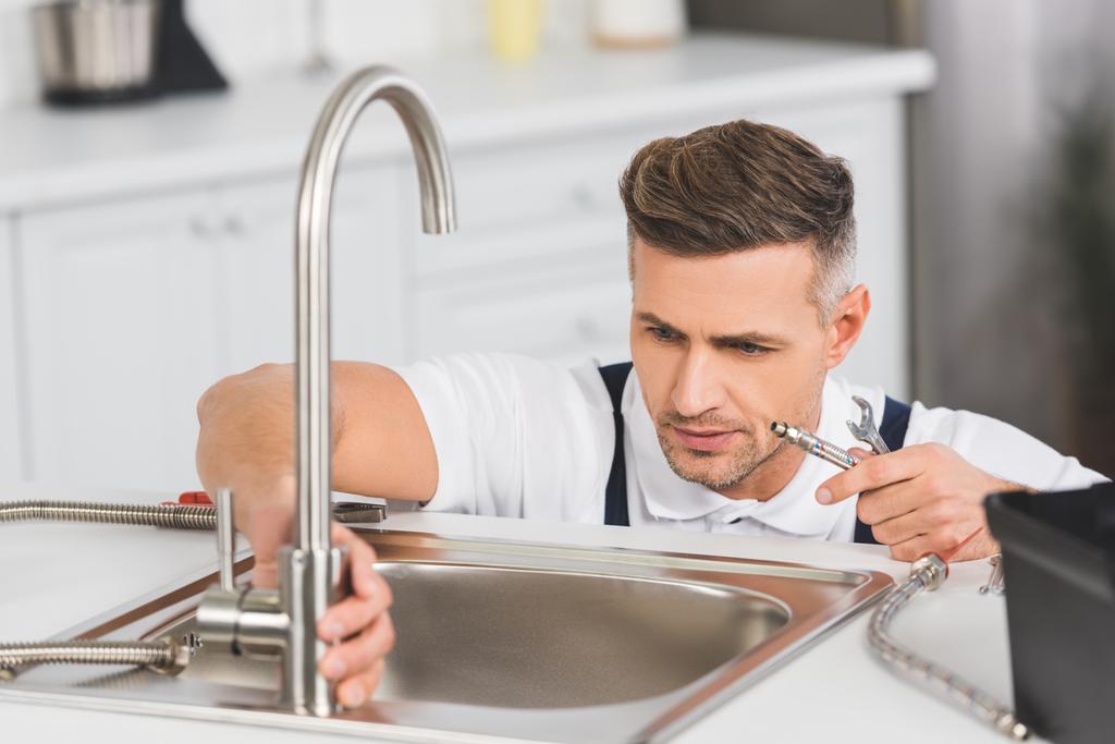 Erwachsener Reparateur hält Rohr und Schraubenschlüssel bei Reparatur des Wasserhahns in der Küche - Foto, Bild