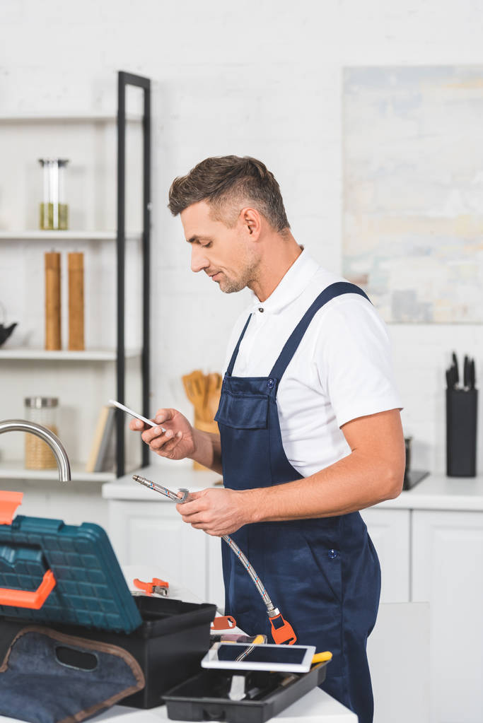 Profil dospělých opravář drží potrubí pro Kuchyně kohoutek opravy a pomocí smartphonu - Fotografie, Obrázek