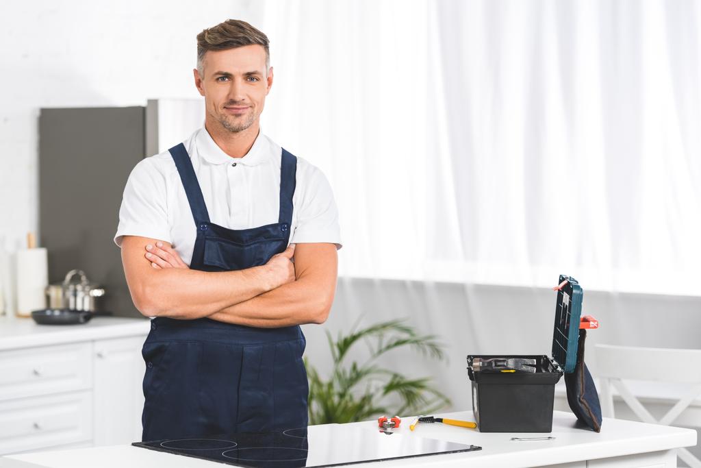 Erwachsener Reparateur steht neben Elektroherd mit gekreuzten Händen in Küche und blickt in Kamera - Foto, Bild