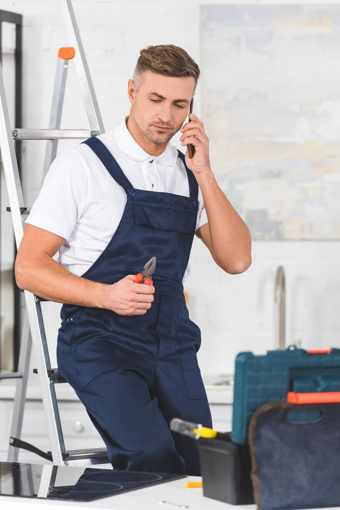 reparador adulto sentado na escada, segurando alicate e falando no smartphone na cozinha
  - Foto, Imagem